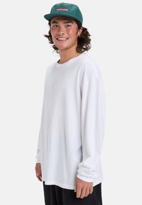 Weißes Langarm-T-Shirt mit lockerer Passform, weichem Material und gesticktem Logo. Getragen mit einer grünen Mütze und schwarzen Hosen.