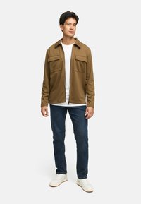 Brun jakke med to frontlommer, båret over en hvit T-skjorte, kombinert med mørkeblå jeans og hvite joggesko. Modell stående foran en ensfarget bakgrunn.