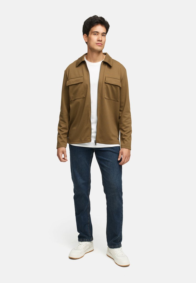 Brun jakke med to frontlommer, båret over en hvit T-skjorte, kombinert med mørkeblå jeans og hvite joggesko. Modell stående foran en ensfarget bakgrunn.