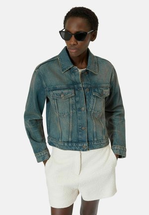 Kobieta w ciemnych okularach przeciwsłonecznych, niebieskiej kurtce dżinsowej i białych teksturowanych szortach, z rękami w kieszeniach, zwrócona lekko w lewo.