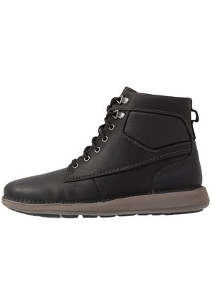 Botte montante en cuir noir avec cheville rembourrée, lacets à œillets métalliques, et semelle en caoutchouc brun clair.