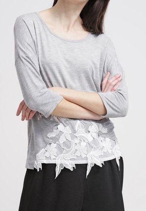 Frau trägt ein hellgraues Langarmoberteil mit weißem floralen Spitzenabschluss und schwarze Hose, Arme vor der Brust verschränkt.