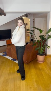 Valge kunstkarusnahast jope lühikese ja keha järgiva lõikega, mis on kombineeritud mustade pükste ja mustade kingadega. Siseruumis, taime ja mööbliesemega.