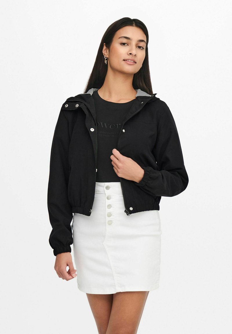 Femme portant une veste noire à fermeture éclair par-dessus un chemisier noir et une jupe blanche à boutons sur le devant, debout devant un fond blanc uni.