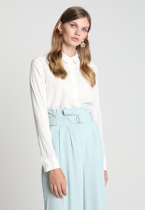 Camicia bianca con bottoni e maniche lunghe, abbinata a pantaloni a vita alta di colore blu chiaro con una cintura dotata di fibbia metallica.