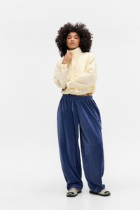 Geel lichtgewicht jack met een hoge kraag, gecombineerd met losse, donkerblauwe corduroy broek. Model draagt instapschoenen.