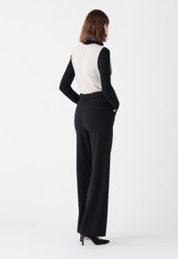 Gilet en tricot crème et noir associé à un pantalon noir taille haute. Le pantalon présente une taille élastique et des poches latérales, associé à des talons noirs.