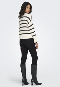 White sweater with black horizontal stripes, paired with black skinny jeans and knee-high black boots. Model is seen from the back.