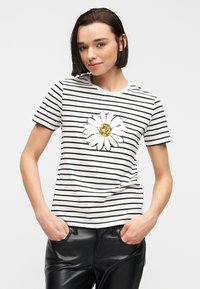 Γυναίκα με κοντά σκούρα μαλλιά, φοράει ένα λευκό και μαύρο ριγέ μπλουζάκι με μεγάλο σχέδιο μαργαρίτας και μαύρο δερμάτινο παντελόνι, με τα χέρια στις τσέπες.