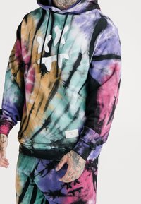 Hoodie w wielokolorowym tie-dye z abstrakcyjnymi wzorami w kolorach fioletowym, turkusowym, czarnym i pomarańczowym, z dużą białą grafiką na klatce piersiowej.