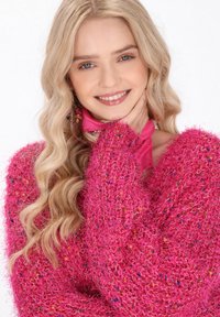 Maglione rosa sfocato con fibre testurizzate vivaci. Collo rotondo, vestibilità rilassata. La modella indossa una sciarpa rosa e orecchini colorati.