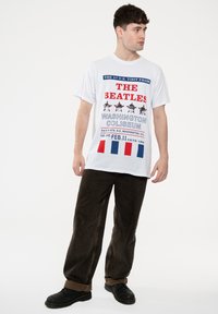 White t-shirt with red and blue print featuring "The Beatles" and concert details. Model wears dark brown pants and black shoes.