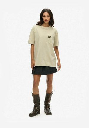 Beżowa, oversize'owa koszulka z kieszonką na piersi, zestawiona z fakturowaną czarną spódnicą plisowaną i skórzanymi, brązowymi butami do kolan z ćwiekami.