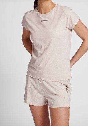 Kobieta ubrana w beżowy t-shirt z krótkim rękawem i szorty z małymi logo "hummel", stojąca z rękami za plecami na jednolitym tle.