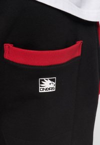 Schwarze Stofftasche mit rotem, geripptem Rand und weißem Logo, das einen stilisierten Vogel und den Text "DNGRS" auf Kleidung zeigt.