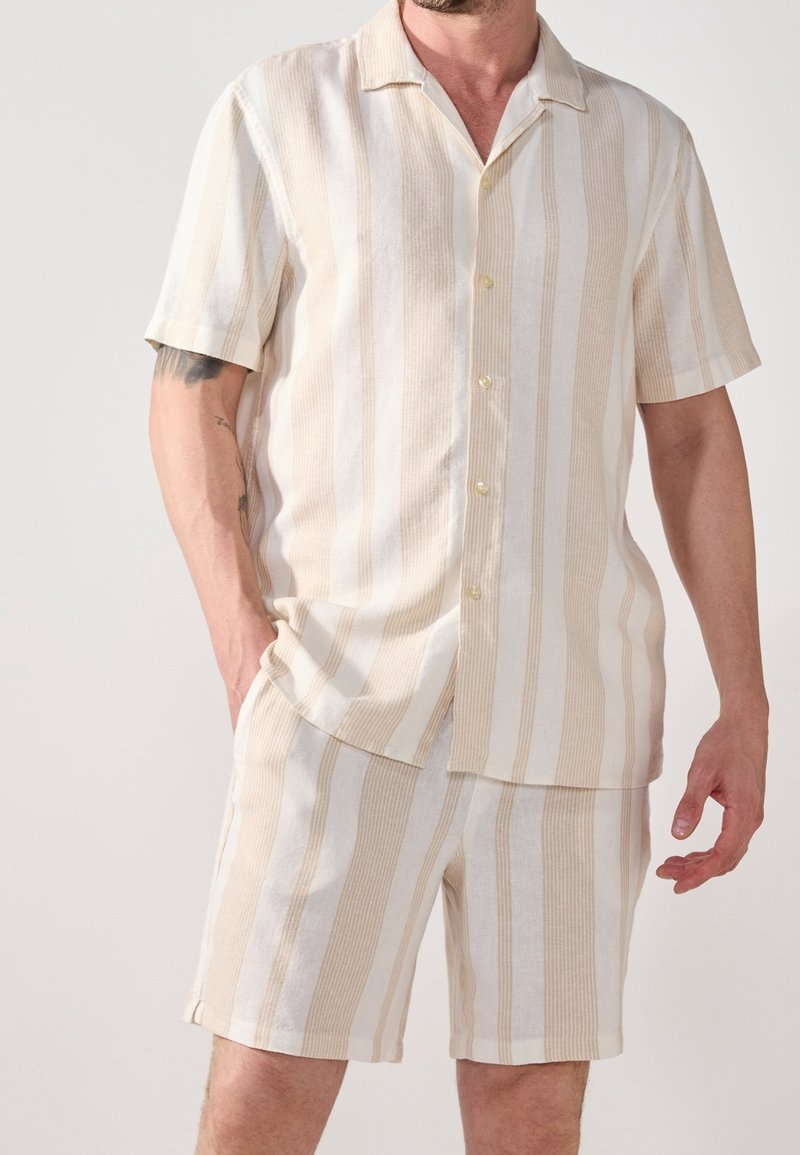 Homme portant une chemise beige et blanche à rayures verticales, à manches courtes boutonnée, et un short assorti, une main dans la poche, sur fond uni.