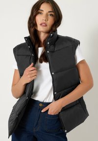 Gilet en duvet noir avec un col haut, fermeture éclair à l'avant et boutons pression. Associé à un t-shirt blanc et un jean taille haute bleu foncé.