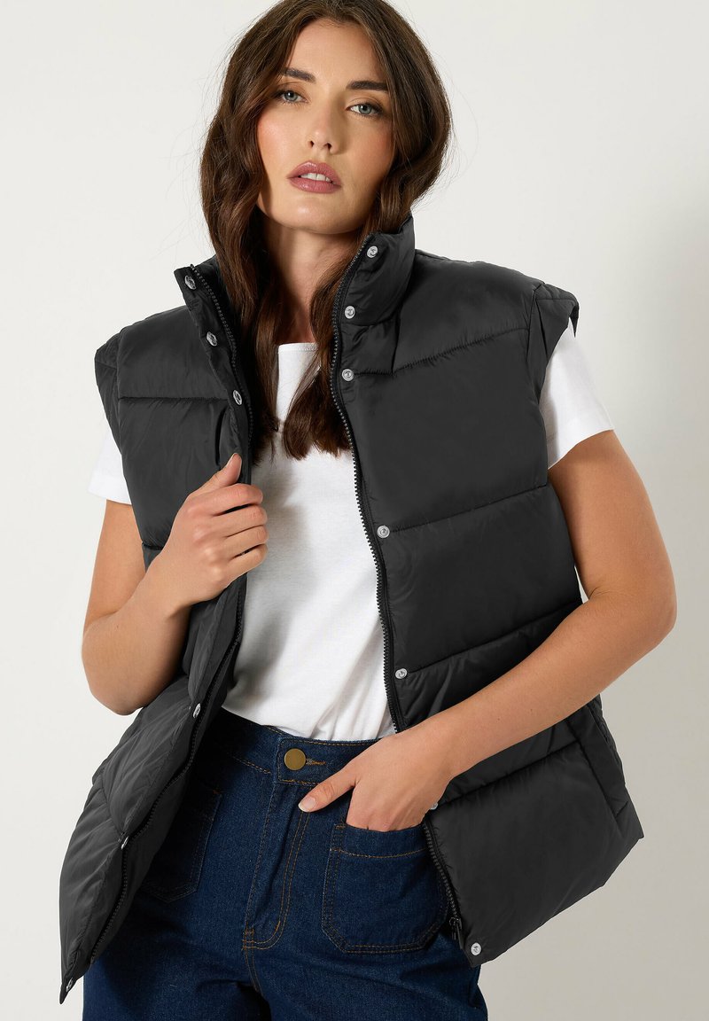 Gilet en duvet noir avec un col haut, fermeture éclair à l'avant et boutons pression. Associé à un t-shirt blanc et un jean taille haute bleu foncé.