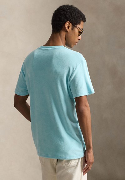 Άνδρας που φοράει ανοιχτό μπλε πουκάμισο με κοντά μανίκια και μπεζ παντελόνι, στέκεται με την πλάτη στραμμένη προς την κάμερα, μπροστά σε απλό μπεζ φόντο.