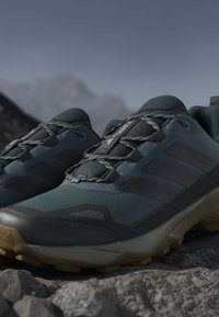 Ciemnozielone buty sportowe z teksturowaną siateczką u góry, gumową podeszwą i szarymi sznurówkami. Posiadają wzmocniony czubek oraz trzy ciemne paski po bokach.