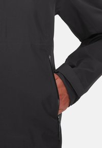 Schwarze Jacke mit einer strukturierten Oberfläche. Verfügt über eine Reißverschlusstasche an der Seite und einen taillierten Bund. Das Material wirkt leicht und langlebig.