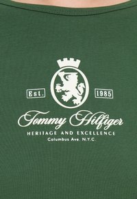 Camiseta de algodón verde con un logotipo blanco que presenta un león y una corona, junto con el texto "Tommy Hilfiger", "Herencia y Excelencia" y "Est. 1985".