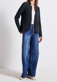 Blazer en maille noire aux bords arrondis, fermeture à un bouton, associé à un jean large bleu et des baskets beiges.