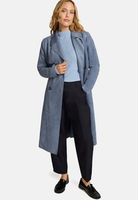 Trench-coat en daim bleu clair avec larges revers, détails à boutons, porté sur un pull côtelé bleu clair, assorti à un pantalon foncé et des mocassins noirs.