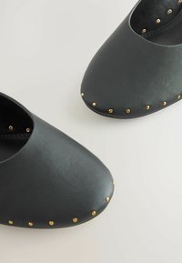Schwarze Schuhe aus Kunstleder mit spitz zulaufender Form und goldfarbenen Nieten entlang des Randes. Glatte Textur mit einem minimalistischen Stil.