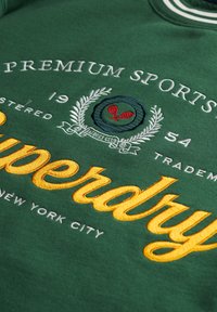 Sudadera verde con cuello a rayas blancas, bordado de "Superdry" en amarillo y un gráfico con raquetas de tenis y diseño de laurel.