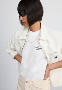 Cream cord jacket with a zip, white t-shirt featuring black text, and pearl necklace; model wearing black sunglasses and light beige pants.