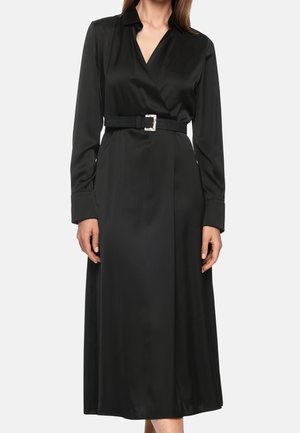 Kobieta w długiej czarnej satynowej sukience kopertowej z długimi rękawami i czarnym paskiem z srebrną klamrą.