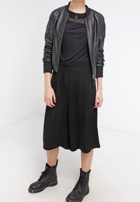 Cappotto bomber in pelle nera sopra una maglietta nera con colletto di perline, abbinato a una gonna midi plissettata nera e stivali da combattimento neri con lacci.