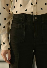 Pantalons en denim noirs avec deux poches avant, associés à un blouse beige à grands pois noirs et poignet boutonné.