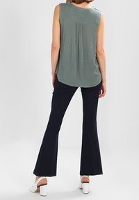 Haut sans manches vert clair avec un détail froncé au dos, associé à un pantalon évasé foncé et des chaussures blanches à talons blocs. Design simple, sans motifs.