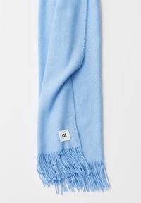 Écharpe bleu clair en tissu doux, avec des bords à franges et une petite étiquette blanche portant un logo noir. Texture lisse sur toute la surface.