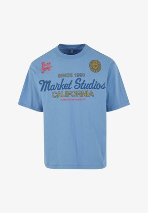 T-shirt en coton bleu clair à manches courtes. Présente un texte en gras en marine et rouge ; les graphiques comprennent "Market Studios" et un logo circulaire.
