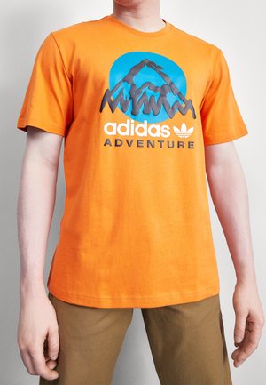 Man die een oranje Adidas Adventure t-shirt draagt met een blauwe cirkel en zwart bergontwerp, gecombineerd met een bruine broek.
