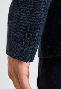 Polsino in tessuto testurizzato blu navy con tre bottoni neri. I bottoni sono rotondi, con quattro piccoli fori, caratterizzati da un design classico e su misura.