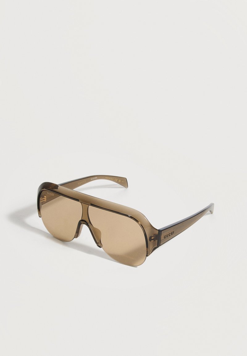 Καφέ αποχρώσεις γυαλιά ηλίου Gucci wraparound με έναν ενιαίο συνεχόμενο φακό και λεπτά, διαφανή μπράτσα σε λευκό φόντο.