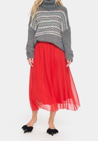 Suéter de cuello alto gris de punto con yugo estampado, combinado con una falda larga de gasa roja y zapatos negros de punta afilada con detalles de lazo.