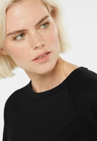 Blonde Frau mit kurzem Haar, die ein schwarzes Shirt trägt und leicht nach links schaut, vor einem schlichten weißen Hintergrund.