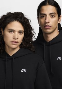 Fekete kapucnis pulóverek puha anyagból, húzózsinórral, kis fehér Nike SB logókkal a mellkasán, két emberen.