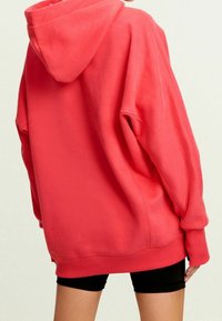 Sweat à capuche oversize corail avec capuche à cordon, poignets et ourlet côtelés. Texture douce avec une coupe décontract ée, porté sur un short noir.