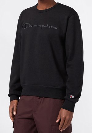 Person trägt ein schwarzes Champion-Sweatshirt mit gesticktem Logo auf Brust und Ärmel, kombiniert mit dunkelbraunen Cargohosen.