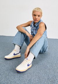 Vita Nike Air Force 1-sneakers med lila detaljer, blå sula och en texturerad överdel. Modellen bär denimoveralls och vita strumpor.