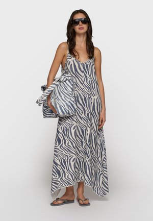 Donna che indossa un vestito lungo senza maniche a righe zebrate e occhiali da sole, tiene una grande borsa a righe zebrate abbinata, in piedi su sandali grigi.