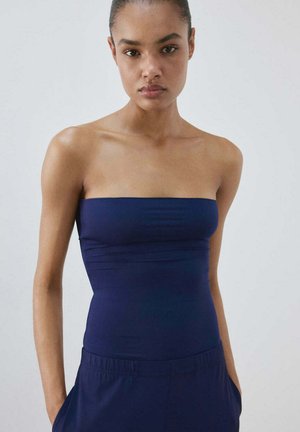 Jeune femme à la peau foncée et aux cheveux attachés, portant un haut bleu marine sans bretelles et un pantalon assorti sur un fond uni.