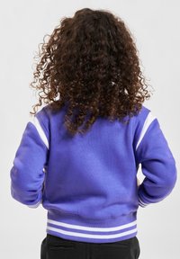 Lila sweatshirt med vita ränder på ärmarna och nederkanten, med en mjuk textur och ribbade muddar. Bakåtriktad vy visas.