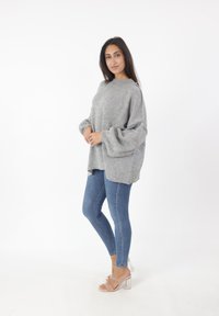 Grijze oversized gebreide trui met verlaagde schouders, gecombineerd met blauwe skinny jeans en doorzichtige hakken, voor een witte achtergrond.